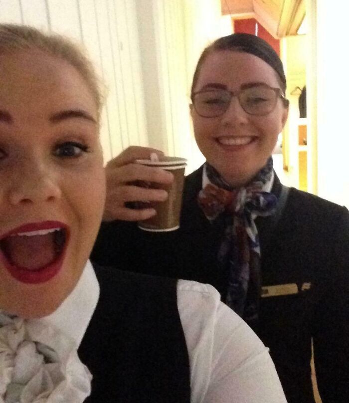 Two women smiling in a candid hotel hallway selfie, one holding a coffee cup. Funny camera roll finds.