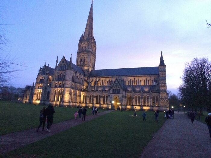 Illuminated church with a tall spire at dusk, surrounded by people enjoying the evening.