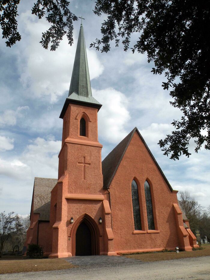 Red brick church with tall spire under a cloudy sky, framed by tree branches.