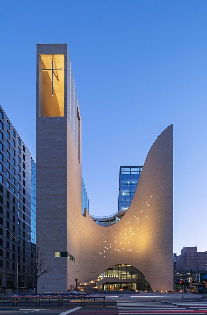 Modern church architecture with a tall cross-lit tower and curved facade, illuminated at dusk.