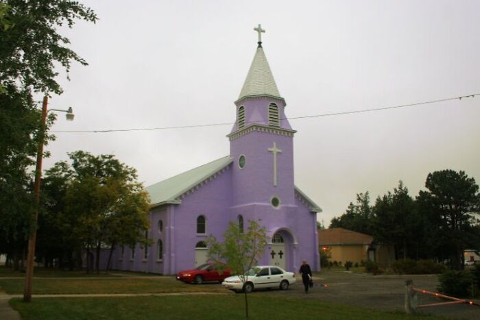 Purple church with a tall spire and cross, surrounded by trees and parked cars, showcasing fascinating architecture.