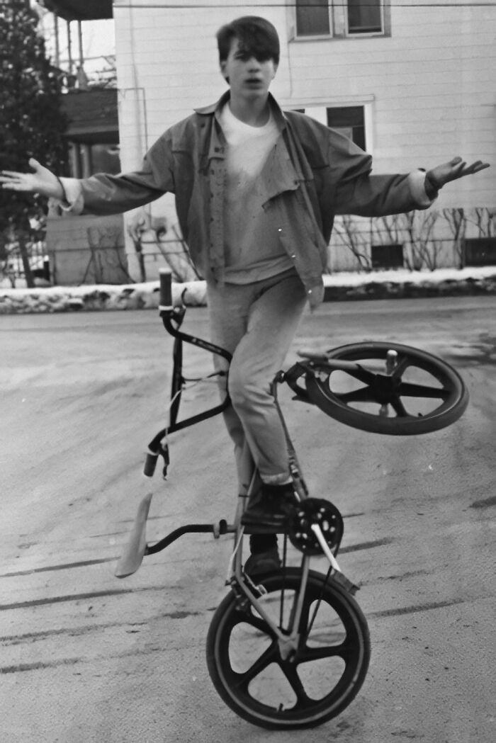 Teen performing BMX trick in street, capturing 80s nostalgia. Wearing casual clothing with hands raised for balance.