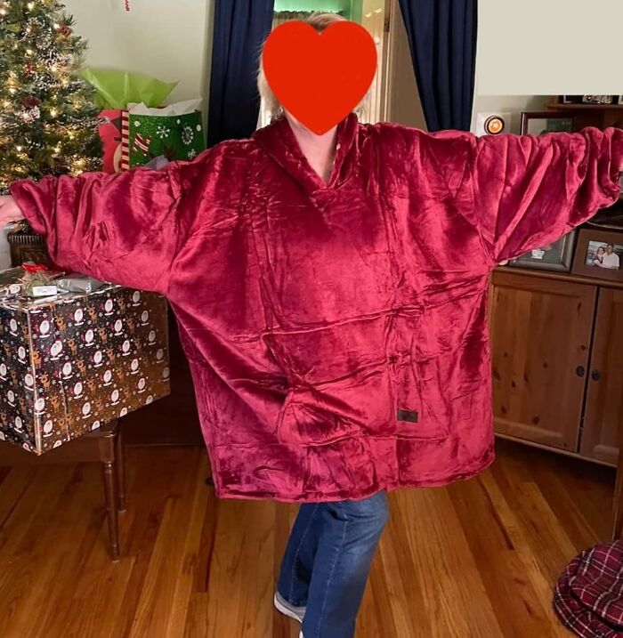 Person wearing a large red cozy blanket sweater inside, standing by a decorated Christmas tree and wrapped gifts.