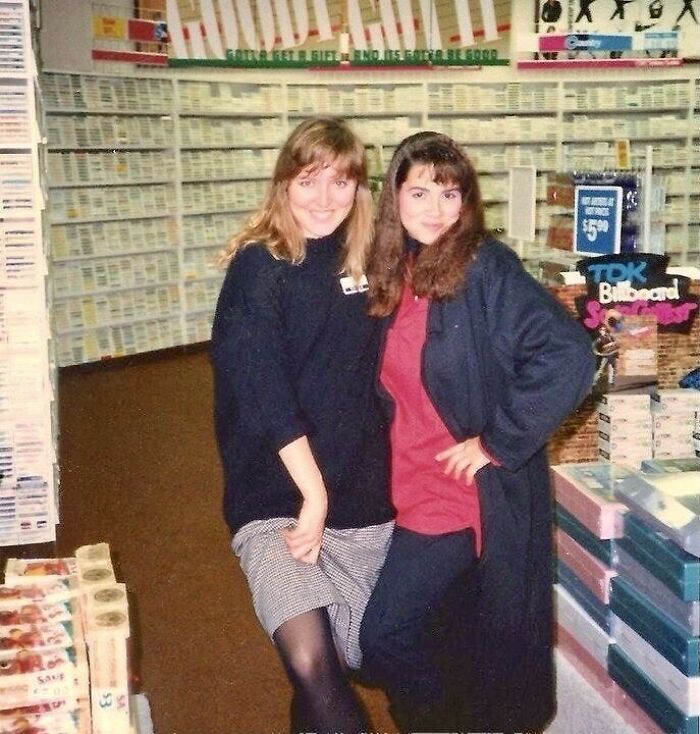 Two women posing in an 80s music store with cassette tapes, capturing 80s nostalgia vibes.