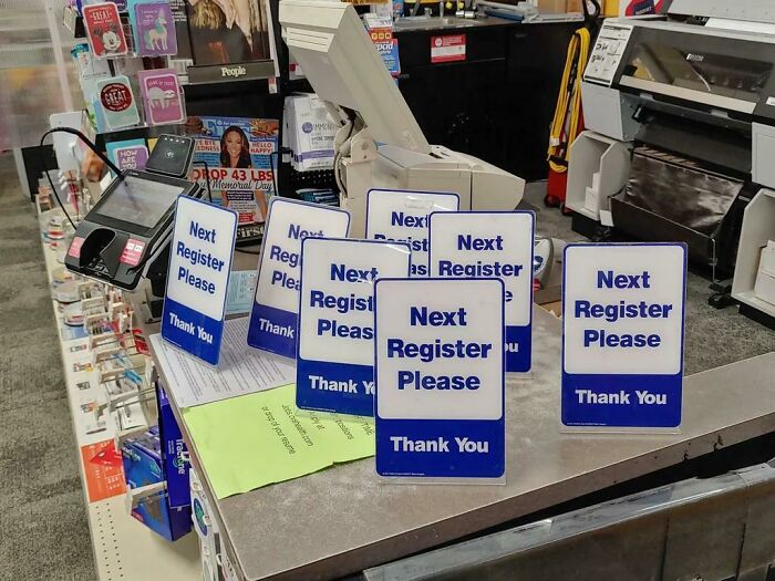 Messy checkout counter in local pharmacy with multiple "Next Register Please" signs.