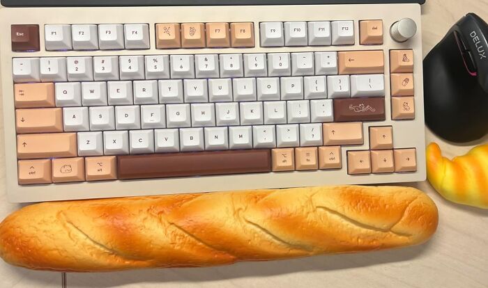 Fun office supplies: a keyboard with playful keycaps and a baguette wrist rest beside a mouse on a desk.