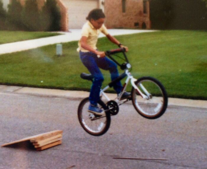 Child on a BMX bike performing a jump over a ramp on a suburban street, capturing 80s nostalgia.