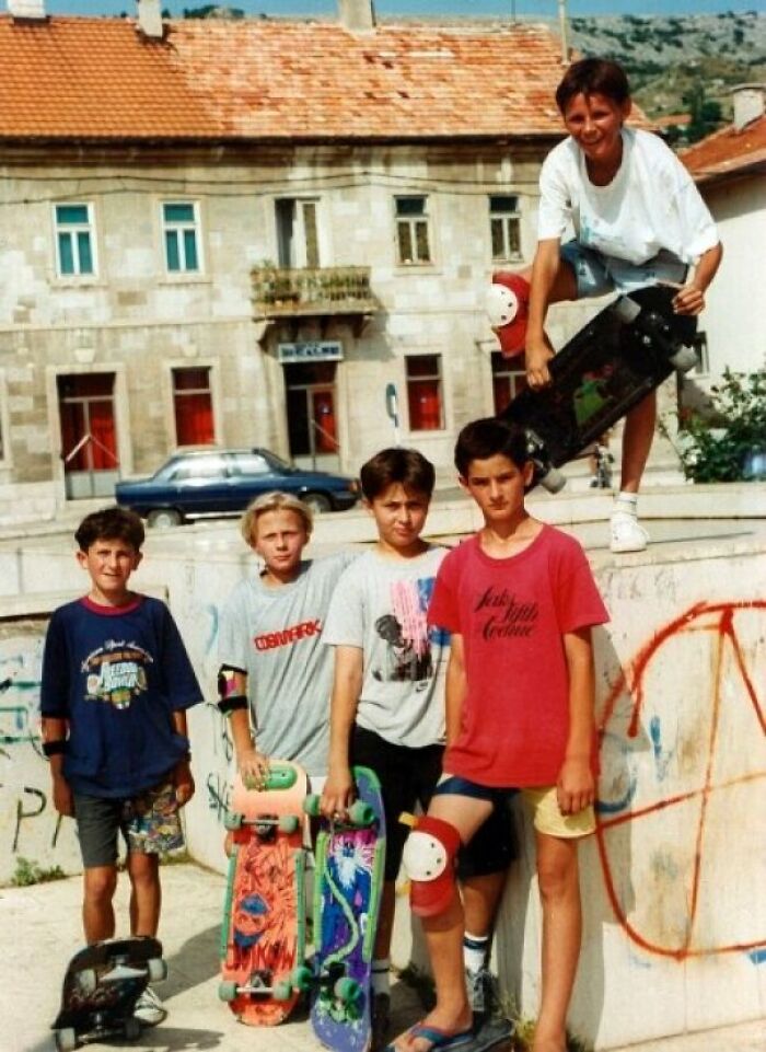 Group of kids in 80s skateboarding gear, holding bright skateboards in front of a graffiti-covered wall.