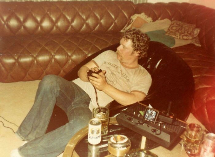 Man enjoying 80s nostalgia, playing video games on a vintage console while relaxing on a beanbag with drinks nearby.