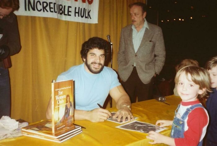 Man signing autographs at a table during an 80s event, with books and kids around.