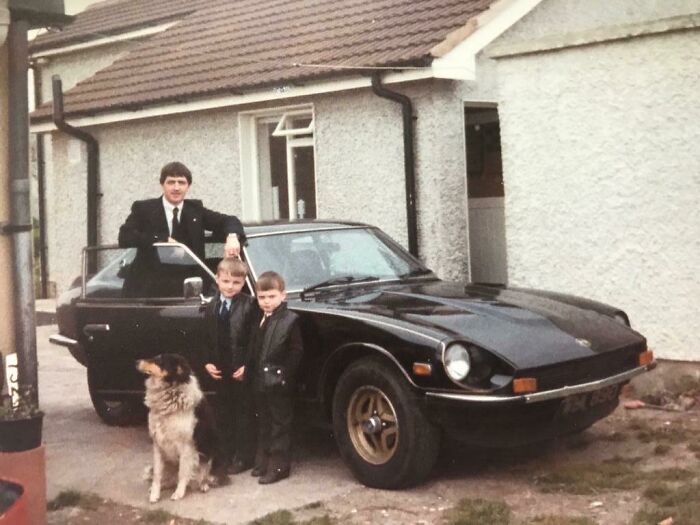 80s nostalgia picture of a man with two boys and a dog standing by a classic black car in front of a house.