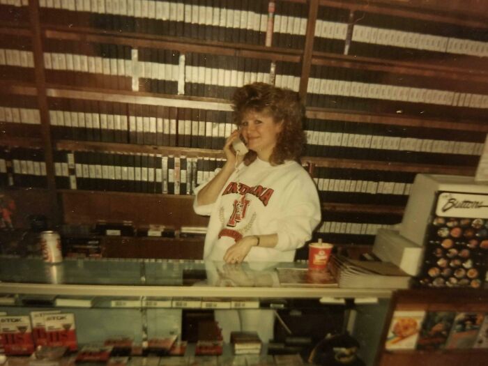 Woman with 80s hairstyle in a video rental store, surrounded by VHS tapes, wearing a college sweatshirt.