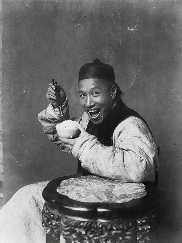 Smiling man in traditional attire enjoying rice with chopsticks, depicting a fascinating historical moment.