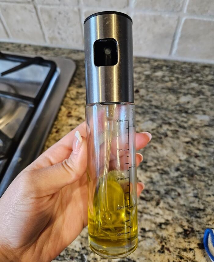 Hand holding olive oil spray bottle in a kitchen, enhancing cooking skills on granite countertop background.