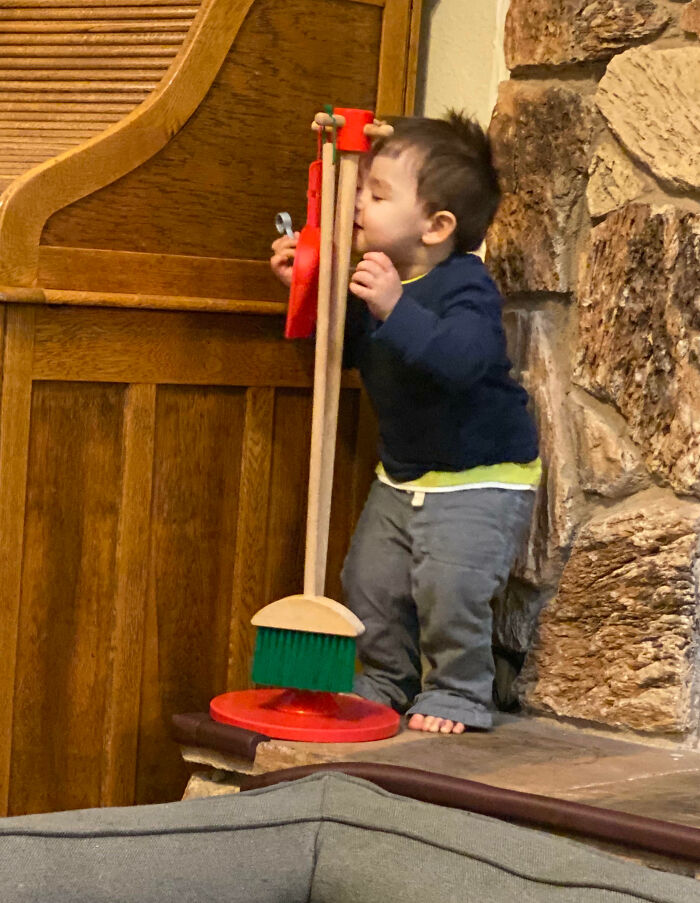 A young child humorously playing with a broom, surrounded by a stone wall and wooden furniture.