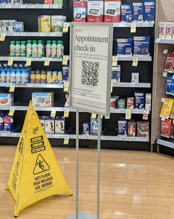 Pharmacy appointment check-in sign with QR code, surrounded by store products and a wet floor caution sign.