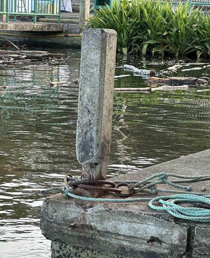 Worn-down concrete post by a riverside with rusted chains and green ropes, showing effects of time and elements.