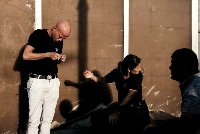 Street photographer captures accidental moment featuring three people and a shadow on a brown wall.