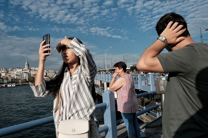 Street photographer captures people on a bridge, each holding their head in various poses against a cityscape background.