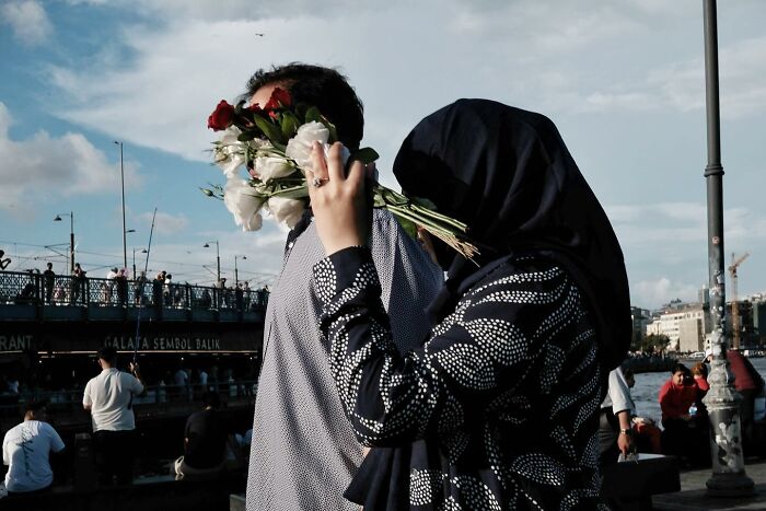 Street photographer captures a humorous scene with a woman holding flowers in front of a man's face by the waterfront.