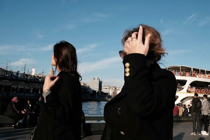 Street photographer captures two women in black coats near a river on a sunny day, with a ferry in the background.