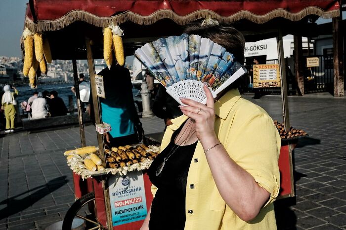 Street photographer captures a woman in yellow hiding her face with a fan near a corn vendor.