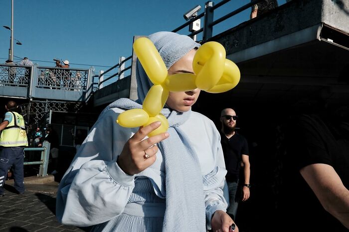 Street photographer captures a woman with a yellow balloon animal obscuring her face, creating a humorous scene.