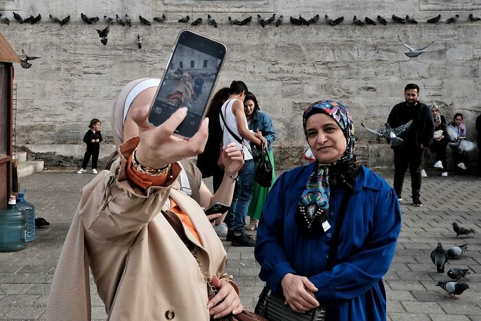 Street photographer captures a funny moment as a woman takes a selfie in a busy area with pigeons and people in the background.