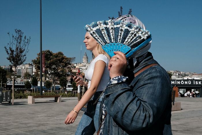 Street photographer captures a humorous scene with one person obscured by a decorative hand fan in a bustling city square.