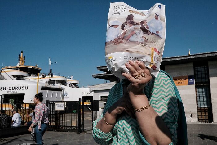 Street photographer captures a funny moment as a person covers their face with a patterned bag near a docked ferry.