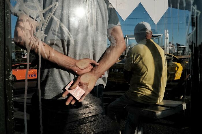 Street photographer captures a humorous scene with a man holding a card behind his back, reflection in a window.