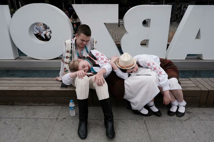 Street photographer captures two women in traditional attire sleeping on a man's shoulder near large sign letters.