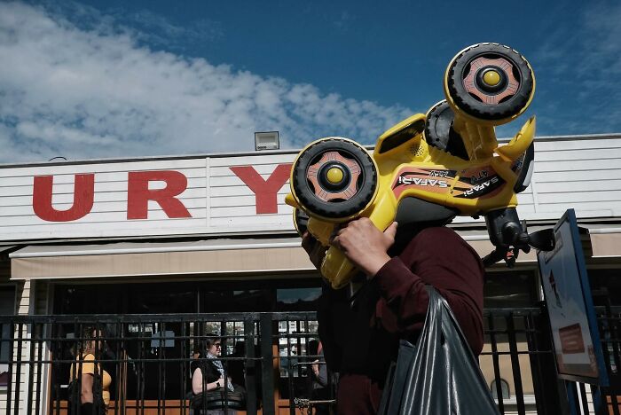 Street photographer captures a funny shot of a person balancing a toy car on their head in front of a shop.