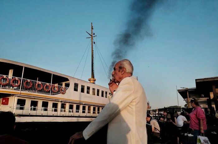 Street photographer captures a man in a white suit smoking, with a ferry and smoke creating an amusing backdrop.