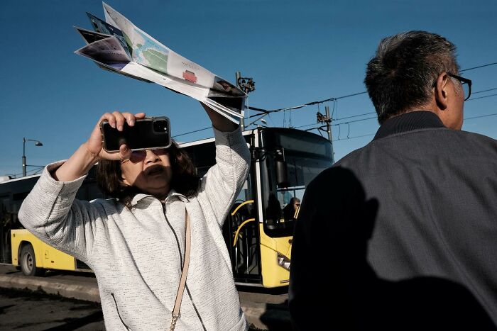 Street photographer captures a woman holding a phone and map in front of a yellow bus on a sunny day.