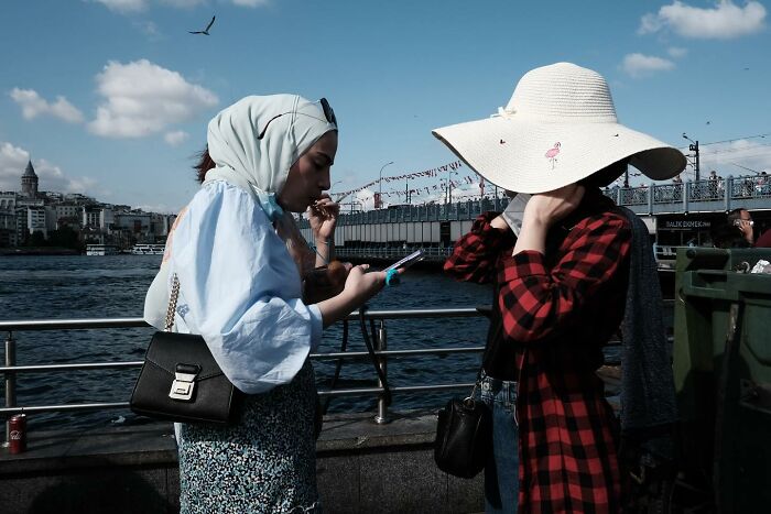 Two women captured by street photographer near water, one wearing a headscarf and the other with a sun hat.