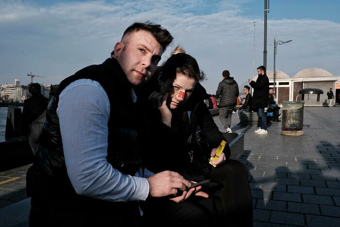 Street photographer captures humorous moment of two people sitting on a bench, cityscape in the background.