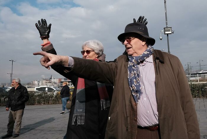 Elderly couple pointing with gloves behind them, creating a humorous accidental image captured by street photographer.