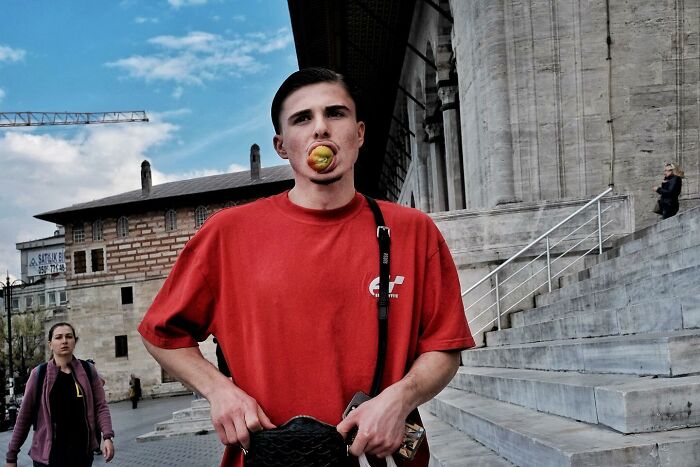 Street photographer captures a man with an apple in his mouth, wearing a red shirt, near grand stone steps.