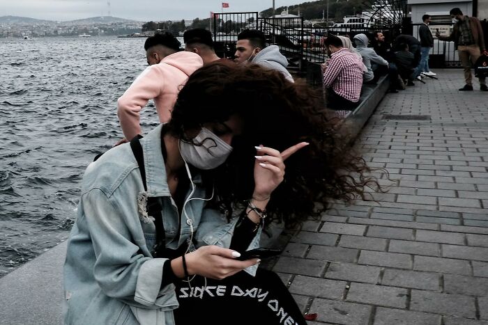 Street photographer captures a woman with curly hair and a mask, sitting by the waterfront, looking at her phone.