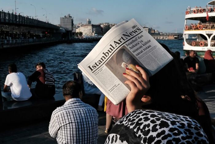 Street photographer captures a person holding a newspaper by the water, showcasing an amusing scene with people in the background.