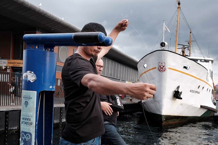 Street photographer captures a funny moment featuring a man appearing to hold a ship with his fishing line.