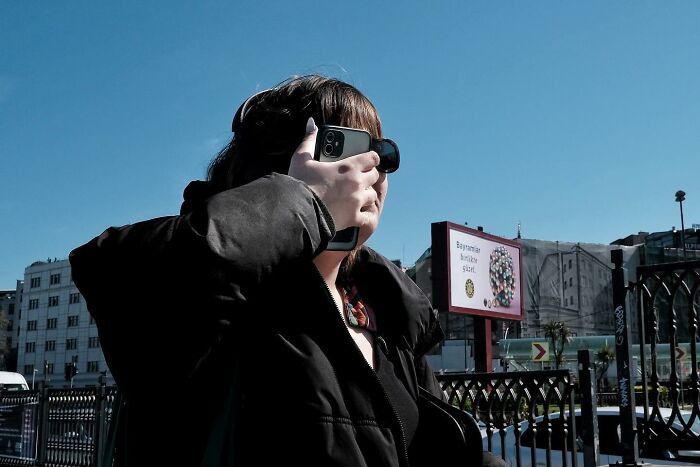 Street photographer captures woman holding phone to face, unintentionally blending with sunglasses, creating a funny scene.