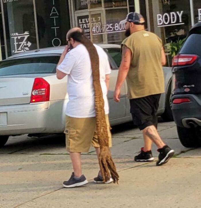 Man with an unusually long dreadlock walking in front of a tattoo shop, highlighting a unique hair accident.