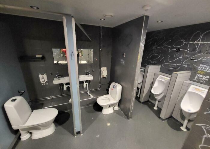 Unfortunate public interior design in a restroom with awkwardly placed toilets, visible urinals, and graffiti-covered walls.