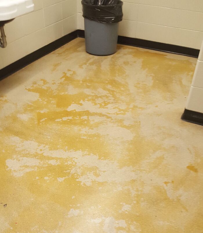 Unfortunate public interior design with stained yellow floor and a gray trash bin in the corner.