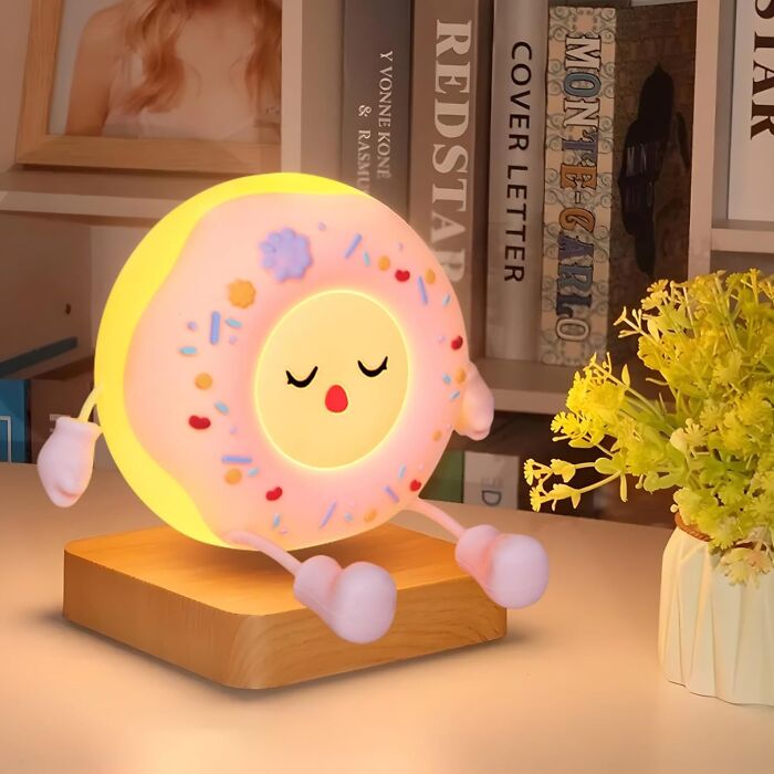 Cute donut-shaped lamp with arms and legs, glowing on a wooden base, next to a plant; Dopamine Decor theme.