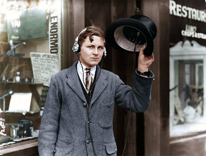 Young man in vintage attire wearing headphones, holding a top hat, showcasing historical inventions.