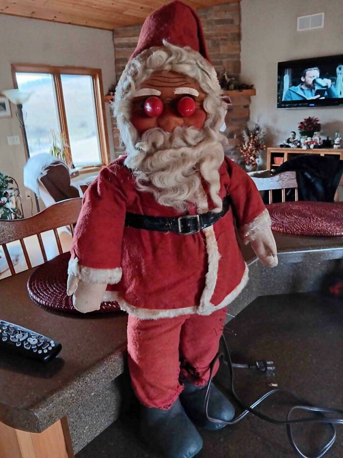 Creepy Santa doll with red eyes and worn suit, standing on a counter in a cozy living room setting.