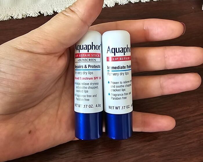 Hand holding two Aquaphor lip repair sticks, essential for addressing unavoidable body issues like dry lips.